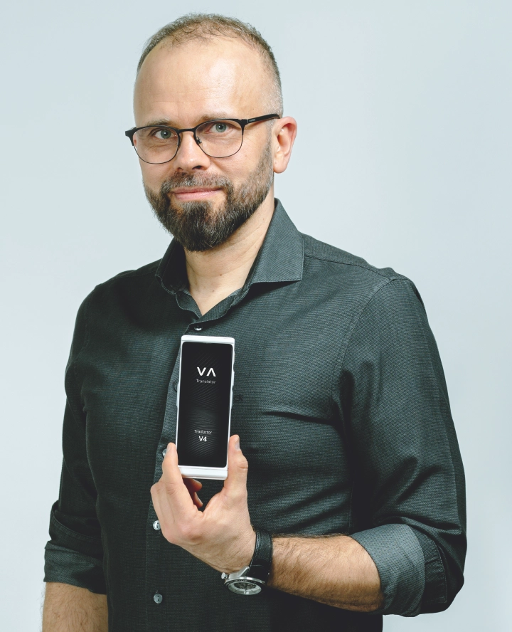  A man with glasses and a beard, wearing a dark shirt, holds up and displays a rectangular device with the letters VA on its screen, smiling slightly at the camera against a plain light background.