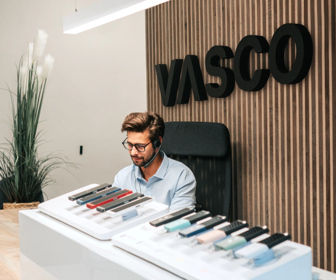 A man from customer service sits at a desk displaying multiple Vasco Translator devices in various colors, with a large 'VASCO' sign on the wooden wall behind him.