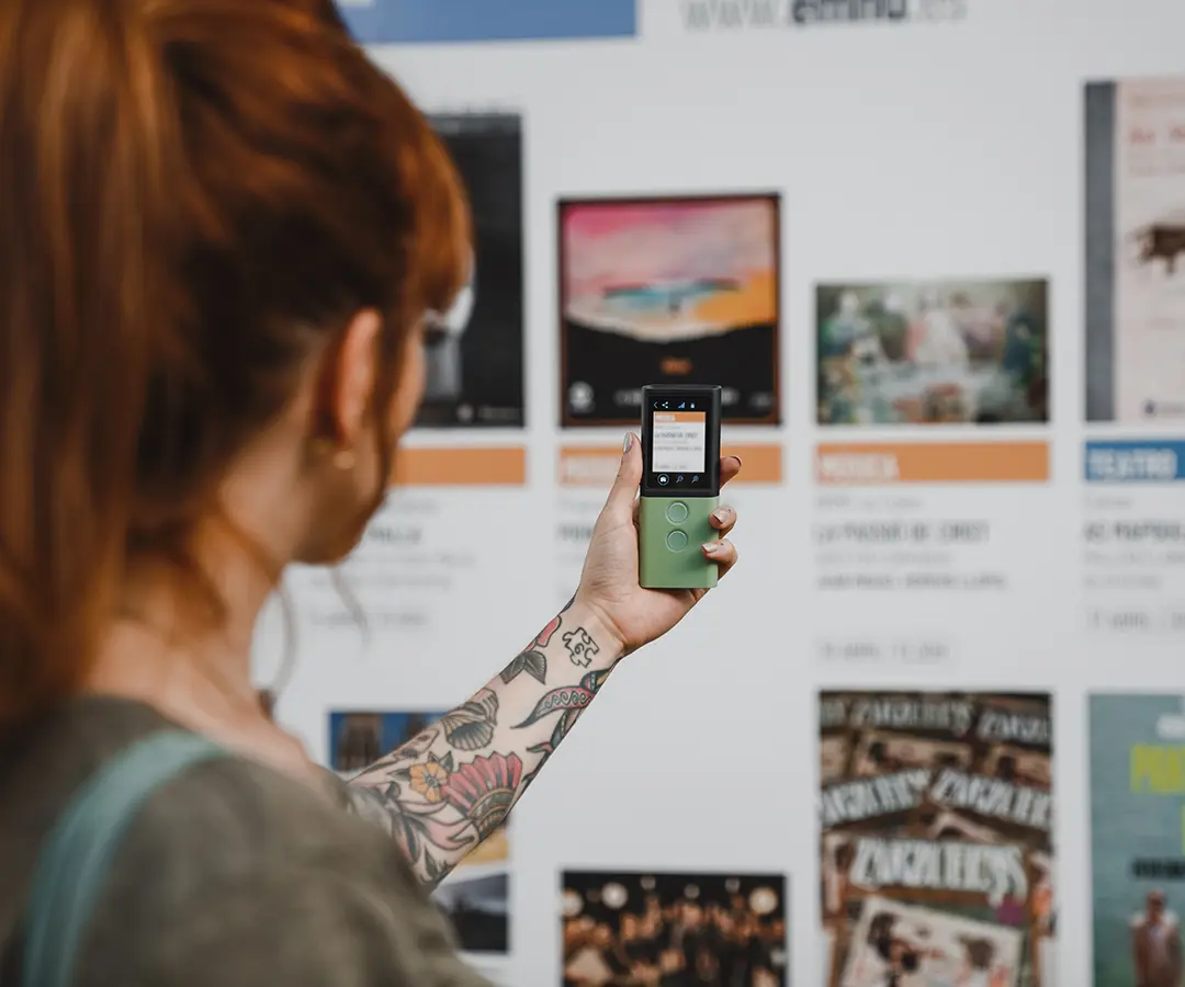A person with red hair and a floral tattoo on their arm holds up a green digital device, pointing it toward a display board with various posters and event information.