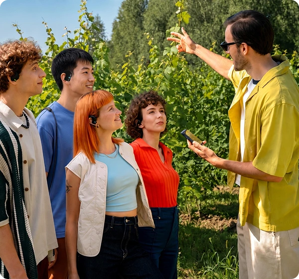 A group of five people outdoors in a sunlit vineyard. On the right, a man wearing a yellow shirt and sunglasses holds a handheld Translator Vasco Q1 device and gestures toward the green grapevines. On the left, four people listen attentively, each wearing a black Vasco Translators E1, showcasing a real-time group conversation feature. 