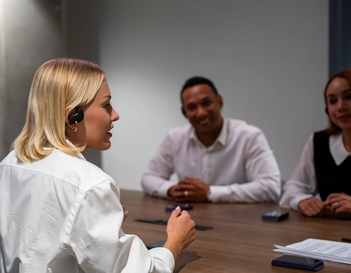 A side profile of a blonde woman in a white blouse speaking during a business meeting at a wooden conference table. She is wearing a black Vasco Translation E1 earbud. In the blurred background, two other professionals are sitting across the table, smiling and listening attentively. 