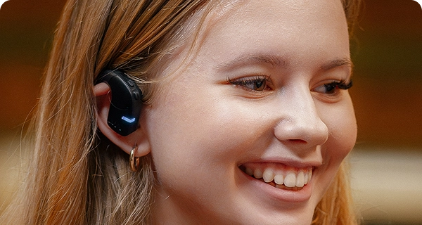 A close-up profile of a smiling young woman wearing a black Vasco Translation E1 earbud. The image highlights the device's hygienic, open-ear design, showing how it sits comfortably on the outside of the ear without entering the ear canal. 