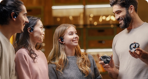 A group of four diverse people showcasing the universal design of Vasco Translation E1earbuds in a warmly lit room. Three individuals are wearing the black earbuds, demonstrating a comfortable fit for different ear shapes, while a smiling bearded man on the right holds two charging cases. 