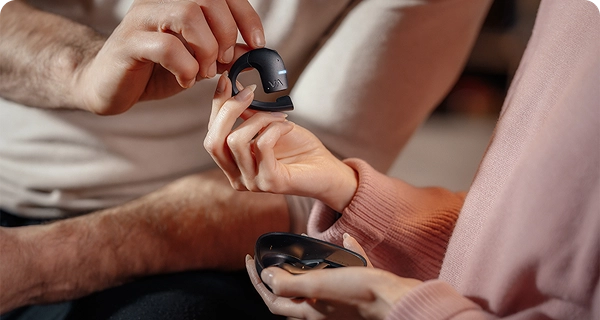 A close-up of a woman's hands gently holding a black Vasco Translation E1 earbud between her fingertips, highlighting its lightweight design. A man's hand is touching the device from above, while she holds an open black charging case in her other hand. 