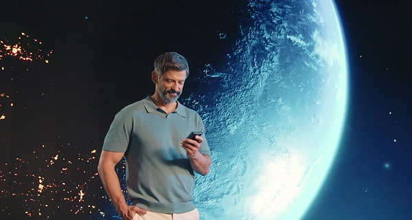 A shot of a middle-aged man with a salt-and-pepper beard and hair, wearing white trousers and a light blue polo shirt with short sleeves. He stands with his right hand in his pocket, looking down at the translator held in his left hand. In the background, a massive, curved display shows a deep space panorama: a large, cold-blue Earth dominates the right side, while glowing golden nebulae and distant city-like lights dot the dark void on the left, amidst scattered stars. The blue light from the Earth projection casts a cool hue onto his side.