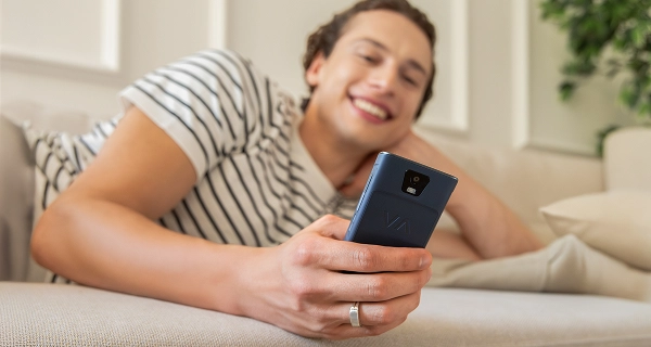 A close-up of a person's hand holding a dark blue pocket-sized device with a VA logo on the back. In the softly blurred background, a smiling young man in a black and white striped t-shirt lies relaxed on a light-coloured sofa. The setting is a bright, modern living room with soft lighting and a hint of indoor greenery.