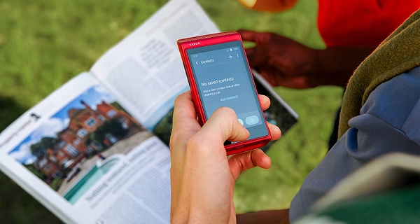 A close-up of a hand holding a red Vasco electronic device over an open travel guidebook or brochure. The display shows a "Contacts" menu with the text "No saved contacts. Add a new contact now or after making a call." alongside an "ADD CONTACT" option. In the blurred background, a green lawn is visible, along with the arm of another person wearing a red shirt.