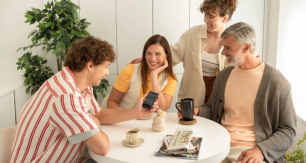 A shot of four people gathered around a small, round white table in a bright, minimalist room. On the left, a man with curly brown hair wearing a red and white striped shirt holds up a dark pocket-sized electronic device, which displays a numeric keypad on its screen. Seated in the middle, a woman in a yellow t-shirt and cream gilet rests her chin on her hand, smiling. Behind her stands a woman with curly hair in a beige shirt, while a grey-haired man with a beard sits on the right in a grey cardigan, holding a black mug. The table is set with coffee cups, newspapers, and a small decorative vase under bright, soft lighting.