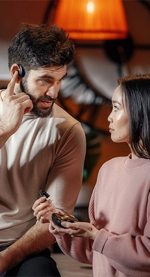 A man wearing the Vasco Translator E1 earpiece, standing next to a woman holding the E1 device, showcasing its powerful speakers and smart microphone.