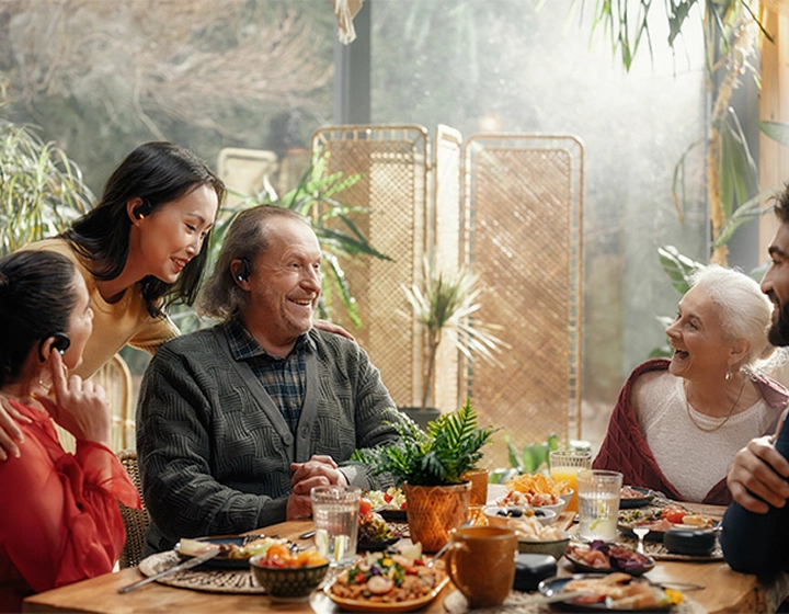 A family sitting around a table, wearing Vasco Translator E1 earpieces, laughing and talking with each other.