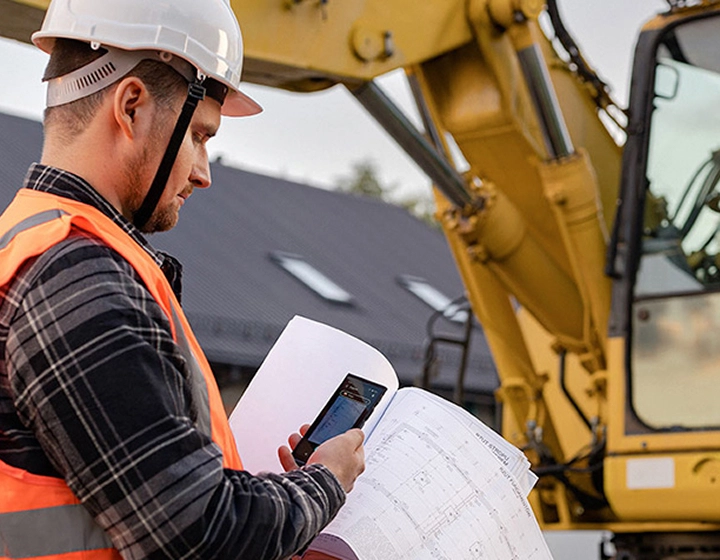 A man wearing a helmet and work clothes taking a photo of construction plans using the Vasco Translator V4, with an excavator visible in the background.