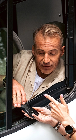 A man sitting in a car, speaking to the Vasco Q1 device that someone is handing to him.