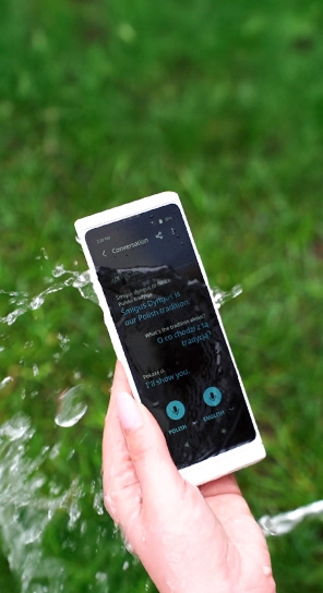 A close-up of a hand holding the Vasco Translator V4 while water splashes over the device, highlighting its durability.