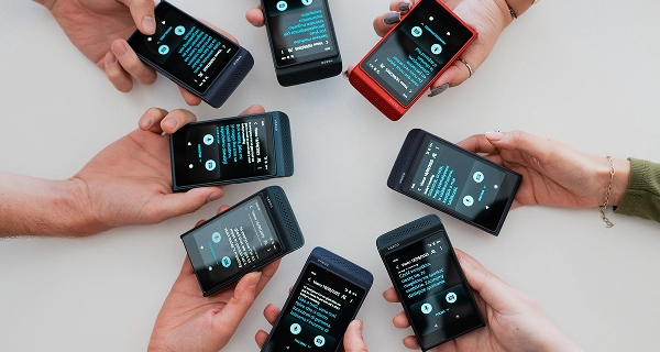A top-down photograph shows seven pairs of hands arranged in a circle, each holding a Vasco Translator Q1 device over a light-colored surface. The devices, in various colors including black, dark blue, and one red, feature bright screens displaying translation interfaces in multiple languages. This visual effectively illustrates the device's ability to facilitate multilingual group conversations.