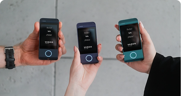 Three people hold up Vasco Translator devices in different colors against a neutral grey background. On the left, a person holds a black device; in the center, a person holds a purple device; and on the right, a person with manicured nails holds a teal device. All three screens are illuminated, displaying the greeting 
