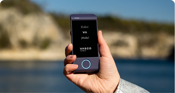 A close-up of a hand holding a purple Vasco electronic translator device against a bright outdoor background. The screen displays the greetings 