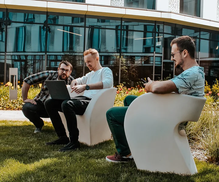 Three men sit on modern white chairs outside a building, working together on a laptop. They appear relaxed and engaged, with one man smiling while looking at his translator. Lush grass and flowers surround them.