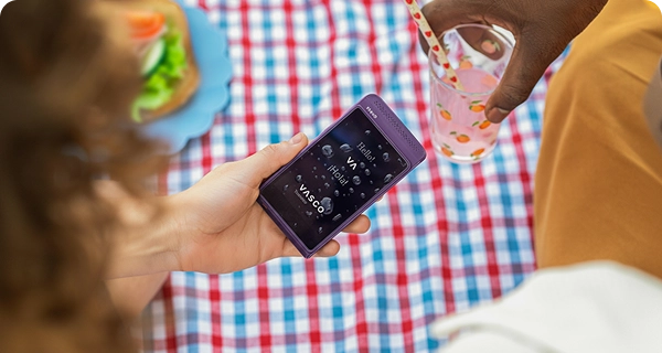 An overhead view of a purple Vasco Translator Q1 being held over a picnic blanket. The device screen is visibly wet, covered in water beads, demonstrating its water resistance feature for outdoor use. In the background, there is a sandwich and a person holding a glass.