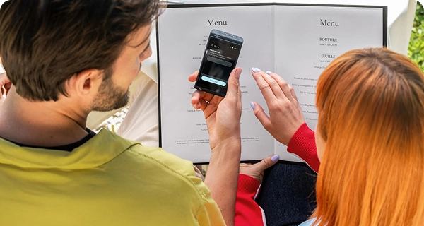 An over-the-shoulder view of a man and a woman with bright orange hair sitting at a restaurant table, looking at a menu. The man holds a black Vasco Translator Q1 displaying a 'Share' screen with an input field to send the translation via email. A white tablecloth and restaurant setting are visible in the background.